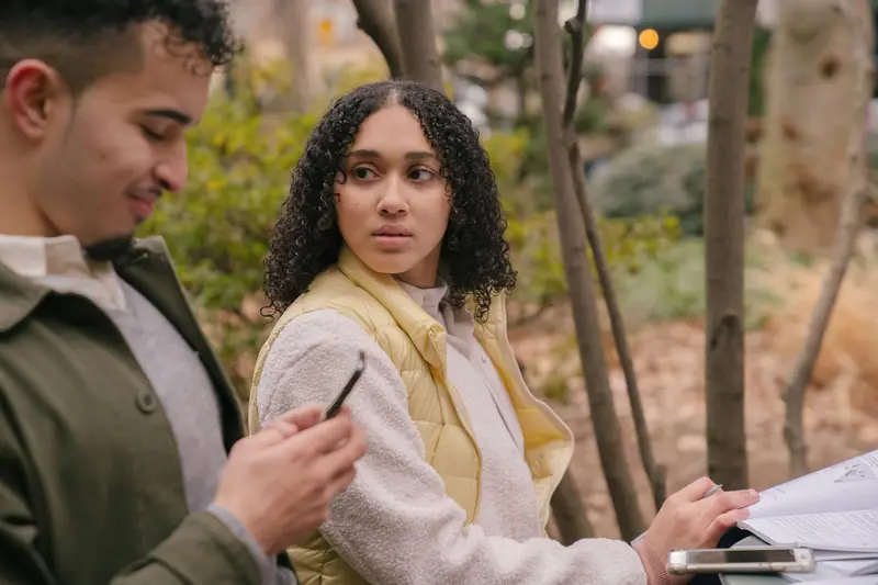 Dos estudiantes en un parque, él con su teléfono y un libro abierto en la mesa.