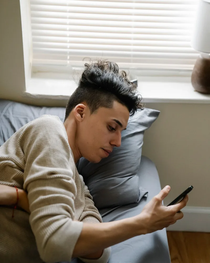 Un joven recostado en su cama revisa su teléfono, a solas y con calma.